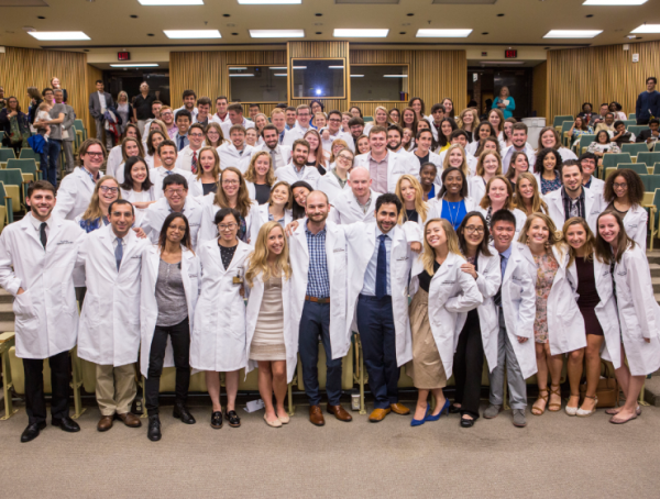 A group of graduate students wearing white lab coats gather in a group for a photo in an auditorium classroom.